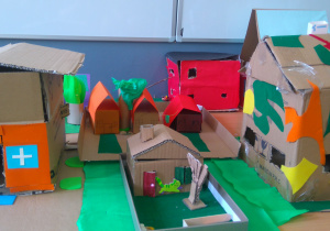 Domki wykonane z tekturowych pudełek oklejonych elementami z kolorowego papieru. Pomiędzy domkami drzewa wykonane z tektury i kolorowego papieru.